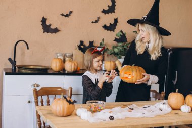 Mother wearing witch costume holding carved pumpkin while daughter making scary gesture with hands in kitchen decorated for Halloween