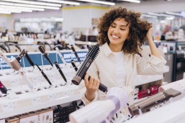 Smiling woman with curly hair touching her locks while selecting a hair dryer in a vibrant appliance store filled with options