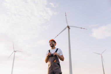 Arab engineer wearing hardhat and work overalls smiling in a wind turbine power plant, renewable energy concept