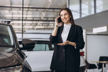 Smiling saleswoman engaging in a phone conversation while presenting a new car to an interested client in a modern car dealership showroom