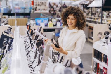 Smiling curly haired woman comparing two hair straighteners while shopping in an electronics store