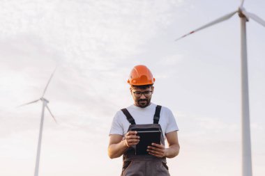 Arab engineer wearing hardhat and work overalls using digital tablet working in wind turbine power plant inspecting sustainable energy production