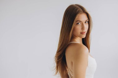 Studio portrait of a young woman with long brown hair wearing a white top, posing against a white background