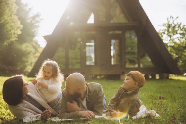 Happy family having fun lying on grass in front of their modern a frame country house, enjoying summer vacation