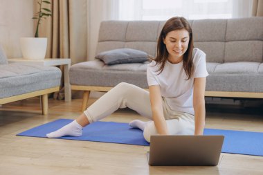 Young woman practicing yoga at home using a laptop placed on a blue mat, following an online class