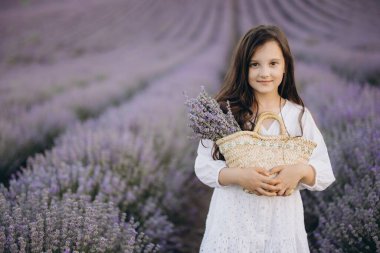 Girl wearing a flowing white dress, holding a wicker basket filled with a vibrant lavender bouquet while surrounded by a blooming lavender field