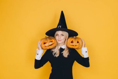 Halloween witch holding two carved pumpkins on her shoulders, smiling slightly on yellow background