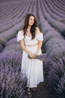 Young woman wearing a flowing white dress, holding a fragrant bouquet of lavender while surrounded by vibrant purple blooms in a serene field
