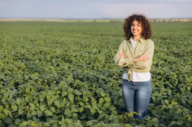 Young agronomist with curly hair standing confidently in a lush soybean field, smiling while enjoying the beauty of sustainable farming