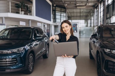 Saleswoman offering car keys and holding a laptop while standing in a car dealership, showcasing professionalism and customer service