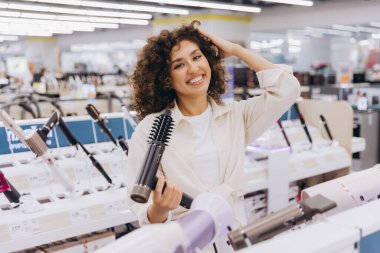 Smiling woman touching her curly hair while browsing through hair dryers in an electronics store, considering her options for purchase