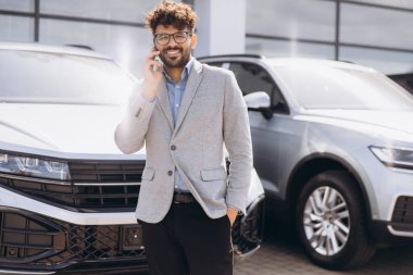 Car salesman engaging in a phone conversation while positioned next to a new vehicle in a bustling car dealership showroom