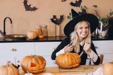 Young blonde woman wearing witch hat carving pumpkins for Halloween party, having fun in her kitchen