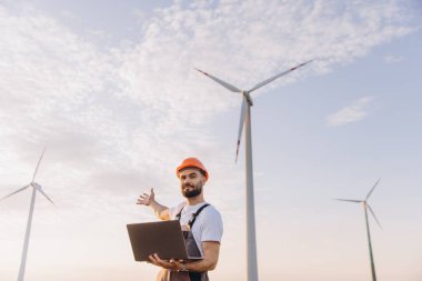 Engineer in coveralls and hardhat is holding a laptop and gesturing towards wind turbines, showcasing sustainable energy generation