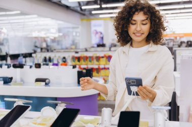 Smiling customer holding smartphone, comparing and choosing mobile phone in consumer electronics store