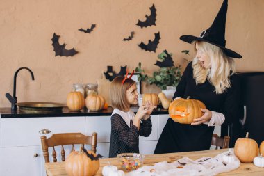 Mom wearing witch costume showing carved pumpkin to her little daughter wearing devil costume in kitchen decorated for Halloween