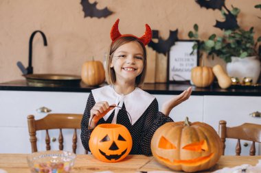 Smiling girl wearing devil costume showing halloween candy basket and carved pumpkin sitting at kitchen table