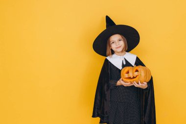Little girl wearing witch costume holding carved pumpkin celebrating Halloween on vibrant yellow backdrop