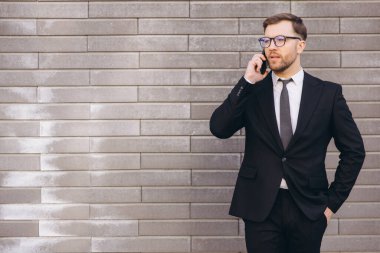 Confident businessman wearing suit and tie talking on smartphone standing near brick wall outdoors