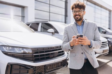 Smiling salesman using a smartphone while standing in front of a lineup of new cars at a busy dealership, showcasing modern automotive options