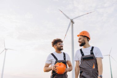 Two engineers discussing next to wind turbines, wearing work overalls and hardhats, in a sustainable energy context