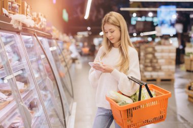 Young woman writing on notepad while carrying shopping basket with fresh vegetables in supermarket