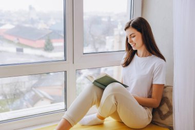 Smiling woman relaxing by the window, immersed in a book and enjoying the peaceful atmosphere of her cozy home during a quiet afternoon