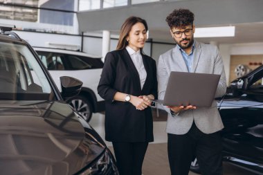 Salesman holding a laptop, assisting a customer in selecting a new car while discussing options in an elegant auto showroom