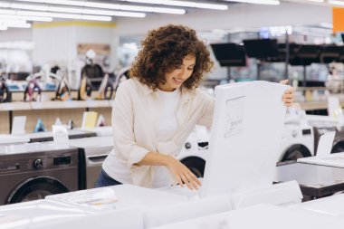 Smiling woman comparing various washing machine models while shopping in an electronics store, enjoying the selection and making a decision