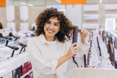 Smiling curly haired woman testing new hair straightener while shopping in consumer electronics store
