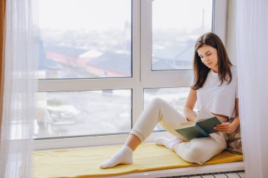 Young woman enjoying a peaceful moment reading a book on a window seat, taking in the city view