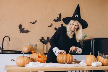 Smiling blonde woman wearing a witch hat joyfully carving pumpkins in a festive kitchen, preparing for a fun Halloween party celebration