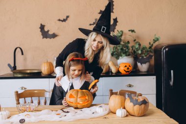 Mother wearing witch hat and daughter carving a jack o' lantern in kitchen decorated for Halloween