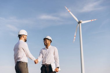 Two engineers shaking hands in front of a wind turbine, celebrating the success of their sustainable energy project