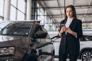 Saleswoman using a mobile phone while standing next to a car in a dealership showroom, engaging with customers and discussing options