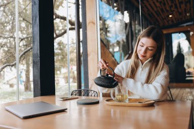 Young woman pouring herbal tea in a transparent cup while working remotely on her laptop in a modern cafe