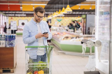 Man using phone and pushing shopping cart with groceries in supermarket