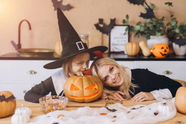 Mother and daughter smiling and having fun while celebrating Halloween at home, wearing costumes and posing with a carved pumpkin