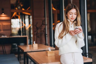 Young blond woman wearing a white sweater is using her smartphone in a cafe, leaning on a table