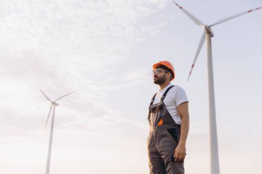 Arab engineer wearing overalls and hardhat inspecting wind turbines in a wind farm at sunset for sustainable energy