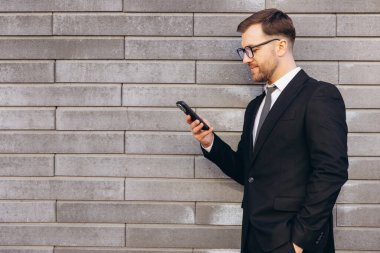 Businessman wearing glasses and suit using smartphone near gray brick wall