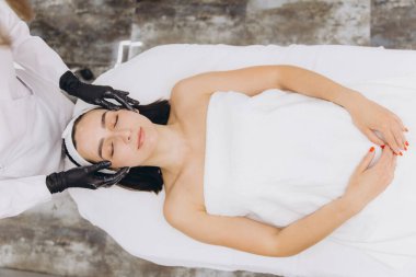 Beautician wearing black gloves massaging face of young woman lying on massage table in beauty salon