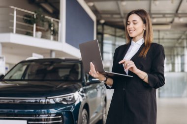 Saleswoman standing next to a new car in a modern dealership, using a laptop to assist customers with online vehicle purchases