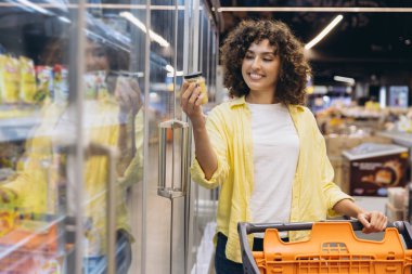 Smiling curly haired woman pushing shopping cart and choosing frozen food in supermarket refrigerator