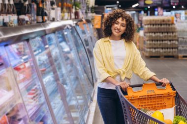 Smiling curly haired woman pushing a shopping cart while walking along refrigerated displays in a supermarket