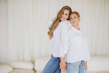 Mother and daughter standing back to back, holding hands and smiling, wearing white shirts and jeans, in a home interior