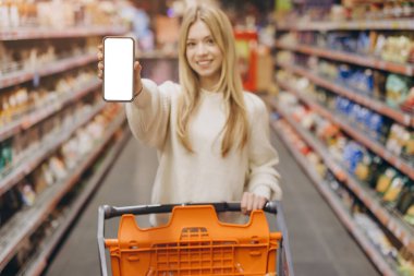 Smiling woman displaying a blank smartphone screen while pushing a shopping cart through the grocery store aisles, enjoying her shopping experience