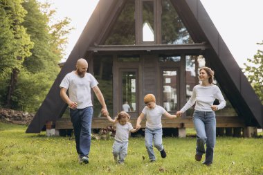 Parents and children holding hands and running on grass in front of their modern a frame country house, enjoying a carefree weekend