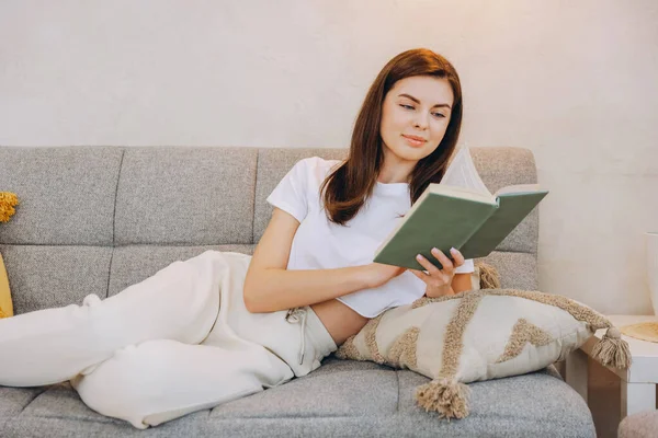 Young woman enjoys a relaxing afternoon at home, comfortably lying on the sofa while engrossed in a captivating book