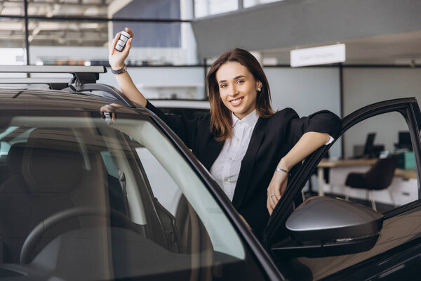 Saleswoman smiling and leaning on a car door, holding car keys, showcasing confidence in a vibrant car dealership showroom